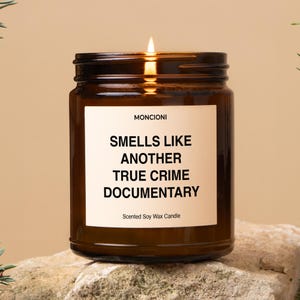 May include: A lit brown glass candle jar with a white label that reads "Smells Like Another True Crime Documentary." The candle is a scented soy wax candle.