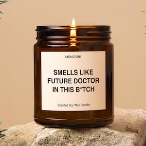 May include: A brown glass candle jar with a white label that reads "Smells Like Future Doctor In This B*tch" and "Scented Soy Wax Candle".