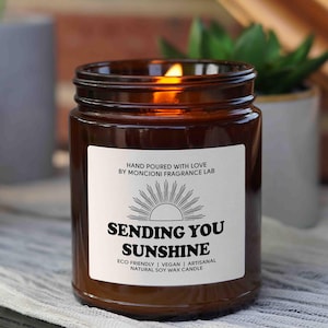 May include: A brown glass candle jar with a white label that reads "HAND POURED WITH LOVE BY MONCIONI FRAGRANCE LAB" and "SENDING YOU SUNSHINE" with a sun graphic. The label also says "ECO FRIENDLY | VEGAN | ARTISANAL NATURAL SOY WAX CANDLE".