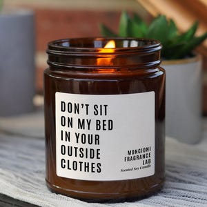 May include: A brown glass candle with a white label that reads "Don't sit on my bed in your outside clothes". The candle is lit and the flame is visible. The label also includes the text "Moncioni Fragrance Lab" and "Scented Soy Candle".