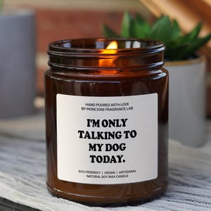May include: A brown glass candle with a white label that reads "I'm only talking to my dog today." The label also includes the text "Hand poured with love by Moncione Fragrance Lab" and "Eco friendly | Vegan | Artisanal Natural Soy Wax Candle".