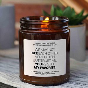 May include: A brown glass candle with a white label that reads "WE MAY NOT SEE EACH OTHER VERY OFTEN. BUT TRUST ME, YOU'RE STILL MY FAVORITE." The candle is hand-poured with love by Moncioni Fragrance Lab and is made with natural soy wax.