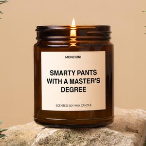 May include: A brown glass candle with a white label that reads "SMARTY PANTS WITH A MASTER'S DEGREE" and "SCENTED SOY WAX CANDLE". The candle is lit and the flame is visible.