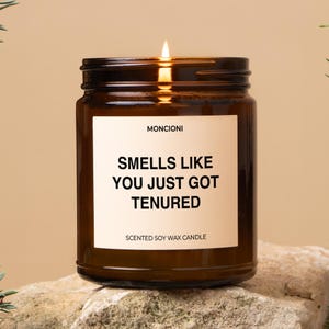 May include: A brown glass candle with a white label that reads "Smells Like You Just Got Tenured" and "Scented Soy Wax Candle".