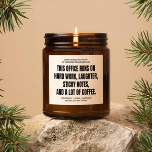 May include: A brown glass candle with a white label that reads "This office runs on hard work, laughter, sticky notes, and a lot of coffee." The candle is lit and sitting on a rock.