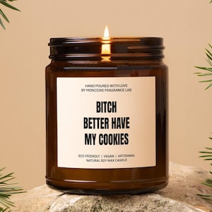 May include: A brown glass candle jar with a white label that reads "HAND POURED WITH LOVE BY MONCIONI FRAGRANCE LAB BITCH BETTER HAVE MY COOKIES ECO FRIENDLY | VEGAN | ARTISANAL NATURAL SOY WAX CANDLE".