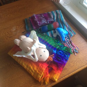 May include: A rainbow colored woven shawl with a white plush doll with a smiling face lying on top. The shawl is on a wooden table with a purple and green striped fabric and a blue and white plastic toy in the background.