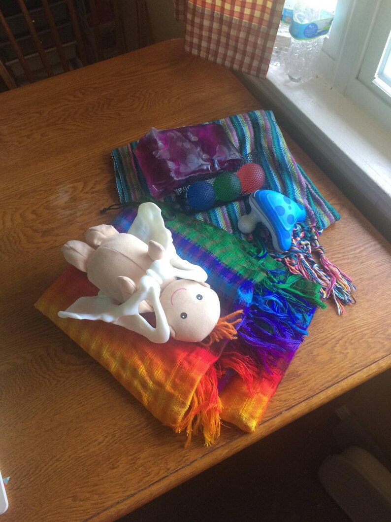 May include: A rainbow colored woven shawl with a white plush doll with a smiling face lying on top. The shawl is on a wooden table with a purple and green striped fabric and a blue and white plastic toy in the background.