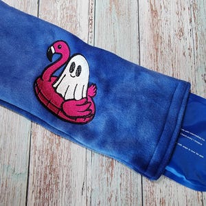 May include: A blue plush heat pack with an embroidered design. The design features a white ghost on a pink flamingo float. A blue gel pack is visible inside the heat pack.