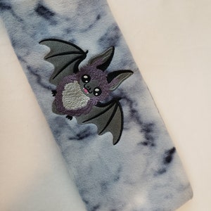 May include: A gray and white tie-dye fabric neck pillow with an embroidered purple bat with gray wings.