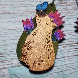 Pu&ograve; includere: Orecchini in legno con un design fantasioso che presenta un orso con una farfalla sulla testa, circondato da fiori rosa e viola su uno sfondo verde.
