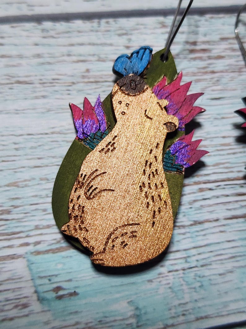 Pu&ograve; includere: Orecchini in legno con un design fantasioso che presenta un orso con una farfalla sulla testa, circondato da fiori rosa e viola su uno sfondo verde.