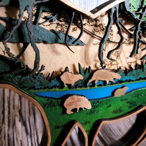 May include: A wooden 3D art piece depicting a forest scene with a river, trees, and animals. The piece is painted in shades of green, brown, and blue.