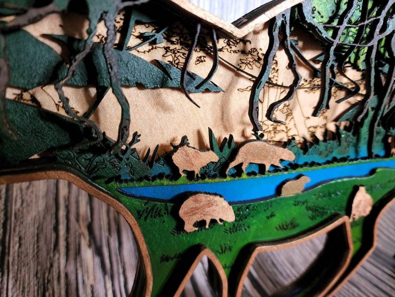 May include: A wooden 3D art piece depicting a forest scene with a river, trees, and animals. The piece is painted in shades of green, brown, and blue.
