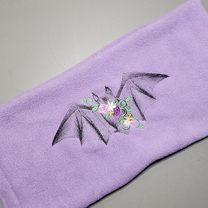 May include: A purple fleece blanket with an embroidered black bat with a floral wreath around its head. The flowers are pink, purple, and yellow.