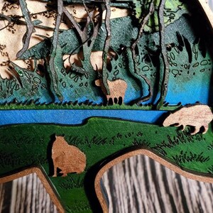 May include: A wooden 3D layered art piece depicting a forest scene with a blue river, green trees, and brown animals. The animals are cut out and layered on top of the background.
