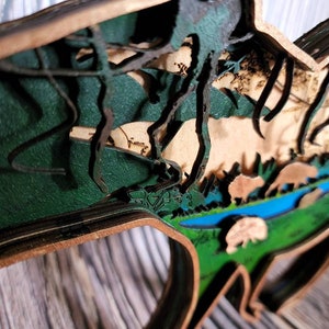 May include: A wooden wall art piece depicting a forest scene with layered wood and green, brown, and blue paint. The scene includes trees, a river, and animals.