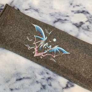 May include: A dark gray, rectangular fabric item with an embroidered bat design. The bat has blue, pink, and white wings, with smaller bats and a moon embroidered around it. The background is a light blue and white cloudy pattern.