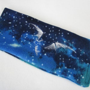 May include: A blue fleece neck warmer with a white embroidered bat flying against a starry night sky background.