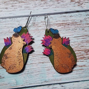 Pu&ograve; includere: Un paio di orecchini in legno con un orso marrone con fiori rosa e viola e una farfalla blu. Gli orecchini sono verdi e hanno un bagliore dorato.