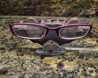 May include: A pair of purple-framed eyeglasses resting on a decorative black and brown holder. The holder has a textured appearance and a small, round base. The glasses have rectangular lenses and a dark purple frame.