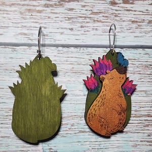 Pu&ograve; includere: Un paio di orecchini in legno con un orso dei cartoni animati con una corona di fiori. L'orso &egrave; marrone con una finitura glitter dorata. I fiori sono rosa, viola e blu. Gli orecchini sono verdi e hanno un gancio argentato.