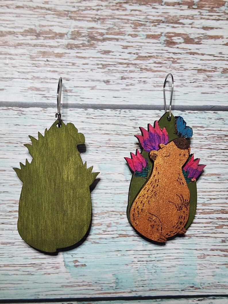Pu&ograve; includere: Un paio di orecchini in legno con un orso dei cartoni animati con una corona di fiori. L'orso &egrave; marrone con una finitura glitter dorata. I fiori sono rosa, viola e blu. Gli orecchini sono verdi e hanno un gancio argentato.