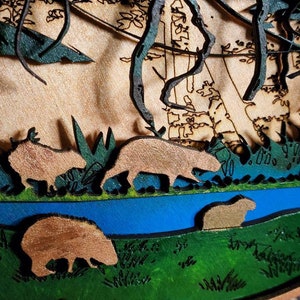 May include: A wooden wall art piece depicting a forest scene with a blue river, green trees, and brown animals. The animals are cut out of the wood and layered to create a 3D effect.
