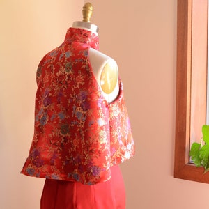 May include: A red and gold brocade top with a mandarin collar and a sleeveless design. The top is on a white dress form.