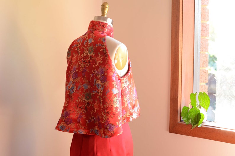 May include: A red and gold brocade top with a mandarin collar and a sleeveless design. The top is on a white dress form.