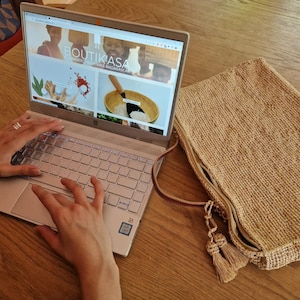 May include: A person is using a silver laptop computer with a website open on the screen. The website is for a company called "Boutikasa" and features images of spices and other ingredients. A brown woven bag is on the table next to the laptop.