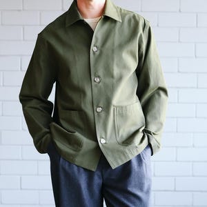 Könnte beinhalten: Eine olivgrüne Jacke mit großen Taschen und silbernen Knöpfen wird mit dunkelblauen Hosen kombiniert. Das Outfit wird durch schwarze Turnschuhe mit weißen Sohlen ergänzt. Die Kleidung ist lässig und für den Alltag geeignet.