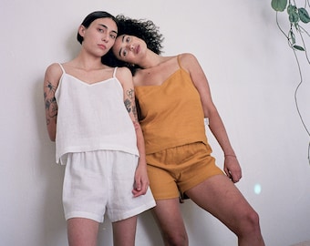 Puede incluir: Un conjunto de camisola y pantalones cortos blancos junto a un conjunto de camisola y pantalones cortos color mostaza. Ambos conjuntos presentan tirantes finos, escotes en V y un corte holgado. Las prendas son de tela texturizada.