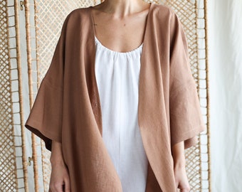 May include: A brown open-front kimono-style jacket with wide sleeves and a straight hem, worn over a white sleeveless jumpsuit featuring a round neckline and wide-leg trousers. The garments are made of a textured fabric with visible weave details.