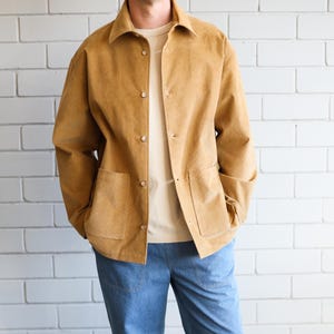 Könnte beinhalten: Eine ockerfarbene Cordjacke mit zwei Vordertaschen und Knopfverschlüssen, kombiniert mit einem cremefarbenen T-Shirt und einer hellblauen Jeanshose. Die Hose ist am Knöchel umgeschlagen, und das Outfit wird durch weiße Leinenschuhe abgerundet.