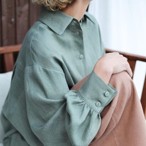 May include: A sage green linen shirt with long sleeves and a button-up front. The shirt has a relaxed fit and features a gathered detail at the cuffs. The model is wearing the shirt with brown linen trousers.
