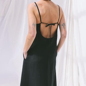 Könnte beinhalten: Ein schwarzes, wadenlanges Kleid mit dünnen Spaghettiträgern, einem tiefen, offenen Rückenausschnitt mit Bindeband und Seitentaschen. Das Kleidungsstück besteht aus einem strukturierten Stoff und fällt gerade bis zum Saum.