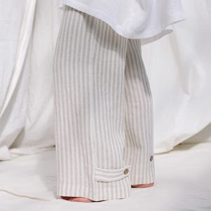 May include: Wide-leg trousers featuring a vertical beige and white striped pattern, paired with a loose-fitting white top. The trousers include a small tab detail at the hem secured with a single brown button, creating a clean and simple aesthetic.