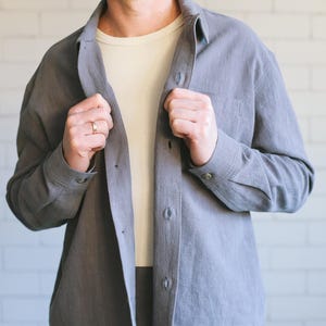 May include: A grey linen shirt and matching trousers set. The shirt features a button-down front, a chest pocket, and long sleeves. The trousers have a pleated front. The outfit is worn over a cream-coloured ribbed undershirt. The background is a white brick wall.