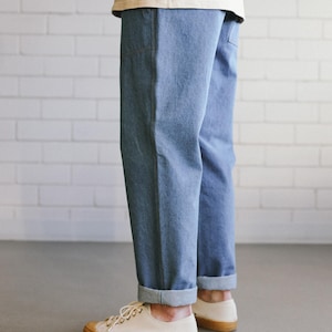 Puede incluir: Pantalones vaqueros azul claro con dobladillos vueltos, combinados con zapatillas de lona color crema con suela de goma marr&oacute;n. La persona lleva una camiseta color crema. El fondo es una pared de ladrillos blancos.