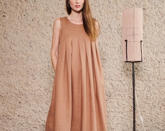 Puede incluir: Un vestido largo sin mangas de color tostado sólido, con escote redondo y pliegues verticales que se extienden desde el pecho hasta el dobladillo. La prenda tiene una silueta holgada y fluida, combinada con zuecos de ante color tostado.