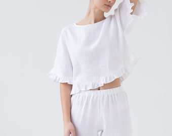 May include: A white two-piece set consisting of a cropped top and matching trousers. The top features a round neckline, elbow-length sleeves with ruffled edges, and a ruffled hemline. The trousers have an elasticated waistband.