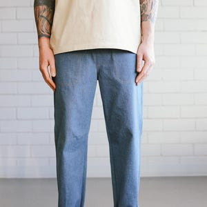 Puede incluir: Pantalones vaqueros azul claro con dobladillos y camiseta beige. Los pantalones tienen dos bolsillos delanteros. La persona lleva zapatillas blancas con suela marr&oacute;n. El fondo es una pared de ladrillos blancos.
