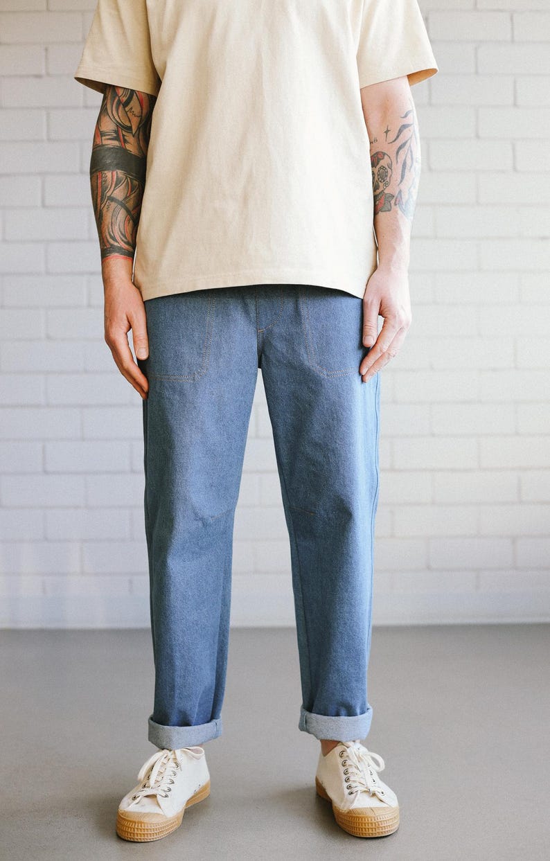 Puede incluir: Pantalones vaqueros azul claro con dobladillos y camiseta beige. Los pantalones tienen dos bolsillos delanteros. La persona lleva zapatillas blancas con suela marr&oacute;n. El fondo es una pared de ladrillos blancos.