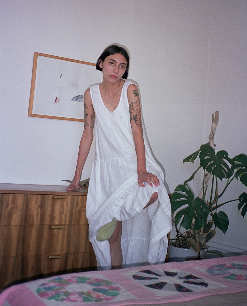 May include: A woman wearing a white sleeveless dress with a ruffled hem. She has tattoos on her arms and is standing in front of a wooden dresser and a potted plant.