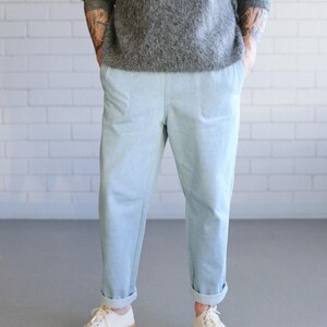 May include: Light blue pants with cuffed hems and a relaxed fit. The pants are paired with white sneakers with tan soles. The person is wearing a gray sweater. The background is a white brick wall.