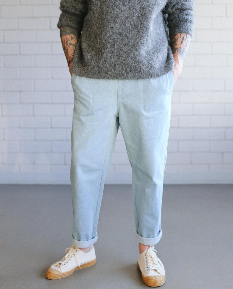 May include: Light blue pants with cuffed hems and a relaxed fit. The pants are paired with white sneakers with tan soles. The person is wearing a gray sweater. The background is a white brick wall.