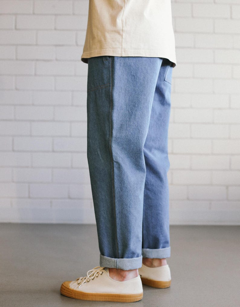 Puede incluir: Pantalones vaqueros azul claro con dobladillos vueltos, combinados con zapatillas de lona color crema con suela de goma marr&oacute;n. La persona lleva una camiseta color crema. El fondo es una pared de ladrillos blancos.