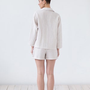 May include: A two-piece pajama set consisting of a long-sleeved button-down shirt with a collar and matching shorts. The garment features a vertical striped pattern in light beige and white, with cuffed hems on the shorts and sleeves.