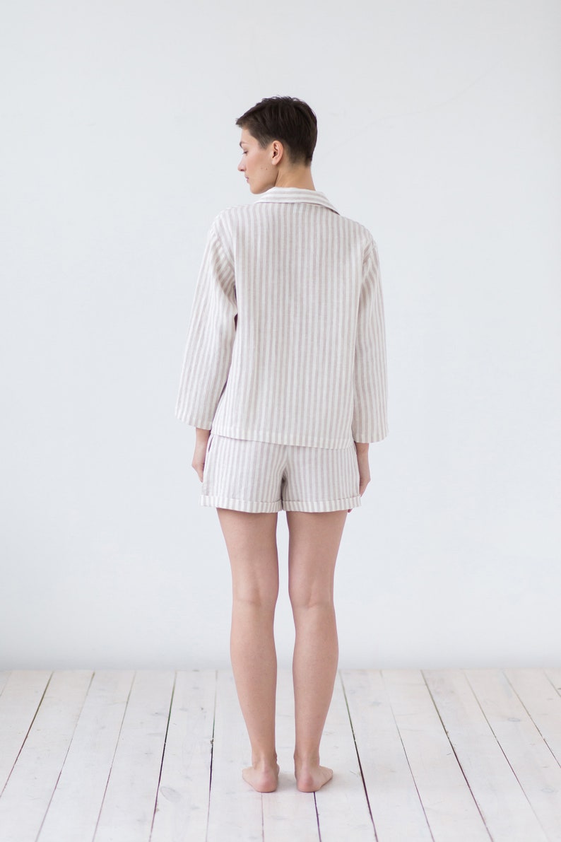 May include: A two-piece pajama set consisting of a long-sleeved button-down shirt with a collar and matching shorts. The garment features a vertical striped pattern in light beige and white, with cuffed hems on the shorts and sleeves.